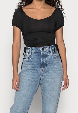 Frau trägt ein schwarzes gerafftes kurzes Top mit kurzen Puffärmeln und hellblauen Jeans mit hoher Taille und Fronttaschen.