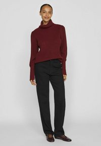 Pull à col roulé en tricot bourgogne avec texture côtelée, associé à un pantalon droit noir et des chaussures marron, sur un fond neutre.