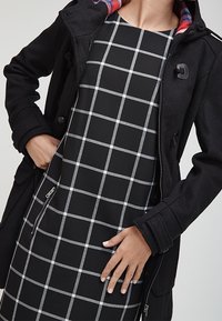 Manteau noir texturé avec doublure à carreaux, porté sur une robe noire avec un motif en grille grise. Détail de fermeture éclair notable sur la robe.