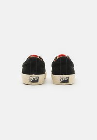 Schwarze Wildledersneaker mit strukturierten Gummisohlen, die rote Akzente an der Ferse und ein prägiertes Logo-Patch auf der Rückseite aufweisen.