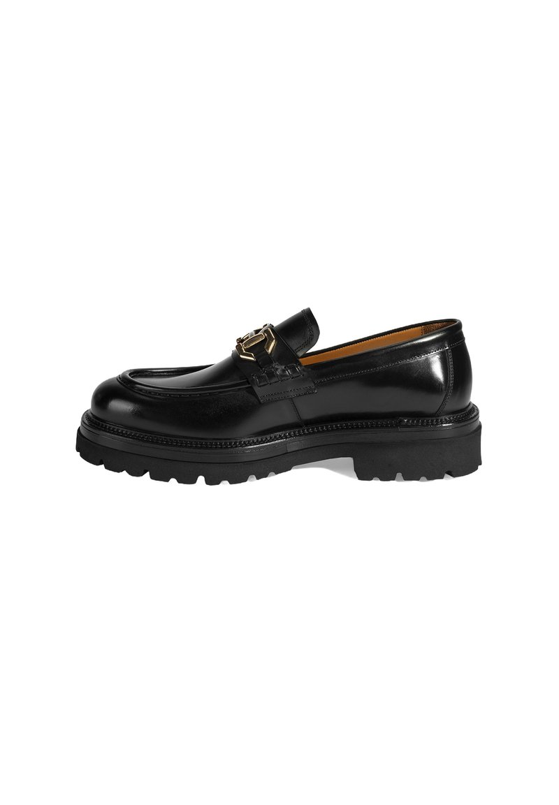 Zwarte leren loafers met een dikke rubberen zool, met een decoratieve gouden hardware-accent over de voorkant en tekstuurelementen.