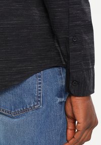Mano di un uomo appoggiata vicino alla tasca posteriore di un jeans blu, indossa una camicia nera a maniche lunghe con polsini abbottonati.