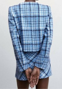 Tummaihoinen henkilö sininen ruudullinen bleiseri ja siihen sopiva hame päällä, seisoo käsivarret ristissä selän takana yksinkertaisen taustan edessä.