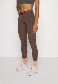 Atletyczna kobieta ubrana w brązowe legginsy Nike Pro, różowe buty Nike i brzoskwiniowe skarpetki Nike, stojąca na tle białego tła.