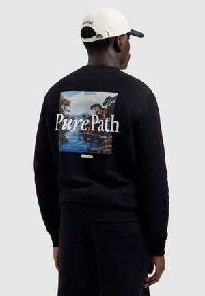 Man draagt zwarte sweatshirt met kleurrijk rivierlandschap en tekst "Pure Path" op de achterkant, witte baseballpet, kijkt weg.