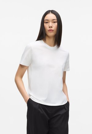 Femme aux cheveux noirs raides portant un t-shirt blanc uni à manches courtes et un pantalon noir, debout les mains dans les poches devant un fond blanc.