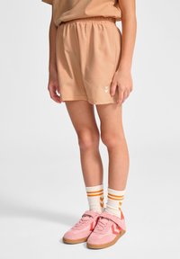 Pfirsichfarbene Shorts mit einem elastischen Bund, ausgestattet mit gestickten Taschen und einem weißen Logo. Kombiniert mit rosa Schuhen und gestreiften weiß-orangen Socken.