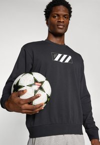 Sort sweatshirt med et tekstureret design, der har et logo og tre hvide striber. Holder en mønstrede fodbold med sorte og grønne accenter.