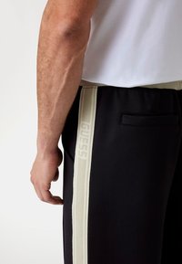 Sorte atletiske joggers med en lys beige sidestrejf og GUESS branding. Glat stoftekstur med sidelomme detaljer.
