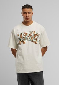 Lyscreme oversized t-shirt med rund hals, med et farverigt grafisk motiv af ænder og teksten "MARHA" trykt på forsiden.