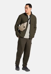 Olive green fuzzy fleece jacket, beige sling bag with zipper, gray t-shirt, cargo pants, and black athletic shoes. Casual outdoor outfit.