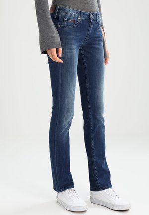 Person wearing fitted dark blue jeans with slight fading and white sneakers, standing against a plain light background.