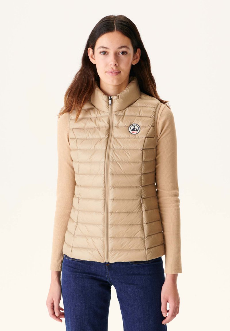 Beige gesteppte Puffer-Weste mit hohem Kragen und Frontreißverschluss, ausgestattet mit einem Logo-Patch auf der linken Brust und seitlichen Reißverschlusstaschen. Getragen über einem beigen Oberteil.