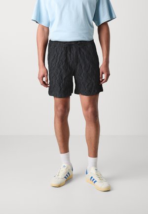 Shorts noirs texturés avec un motif ondulé, taille à cordon de serrage, associés à une chemise à manches courtes bleue et des baskets de couleur claire.