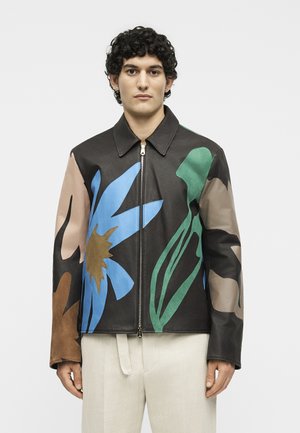 Muškarac s kovrčavom kosom nosi crnu kožnu jaknu s velikim plavim, smeđim i zelenim cvjetnim uzorcima i bež hlače, stoji ispred bijele pozadine.