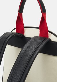 Sangles en cuir noir avec finition texturée, accents en tissu rouge et corps en toile crème. Présente une structure robuste et des couleurs contrastantes.