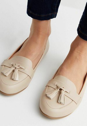 Beige leather loafers with tassel accents, rounded toe, and smooth texture. The design features a stitched trim and flat sole.