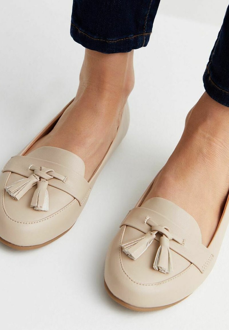 Beige leather loafers with tassel accents, rounded toe, and smooth texture. The design features a stitched trim and flat sole.
