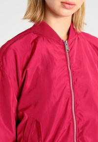 Fuksjowa kurtka bombowca o gładkiej, błyszczącej fakturze; posiada prążkowany kołnierz, zamek błyskawiczny oraz kieszenie boczne.