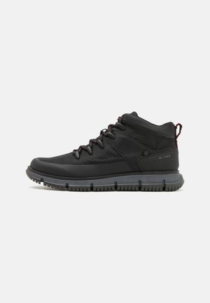 Schwarze Mid-Top-Schuhe aus Mesh- und Synthesematerialien, mit strukturierter Oberfläche, roten Akzenten an den Schnürsenkeln und einer flexiblen grauen Gummisohle.