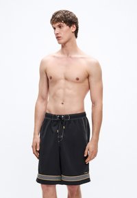 Μαύρο μαγιό σορτς με κορδόνι στη μέση, κίτρινες αντιθέσεις και λευκά ραφές. Σχεδίαση μέχρι το γόνατο με πλαϊνές τσέπες.