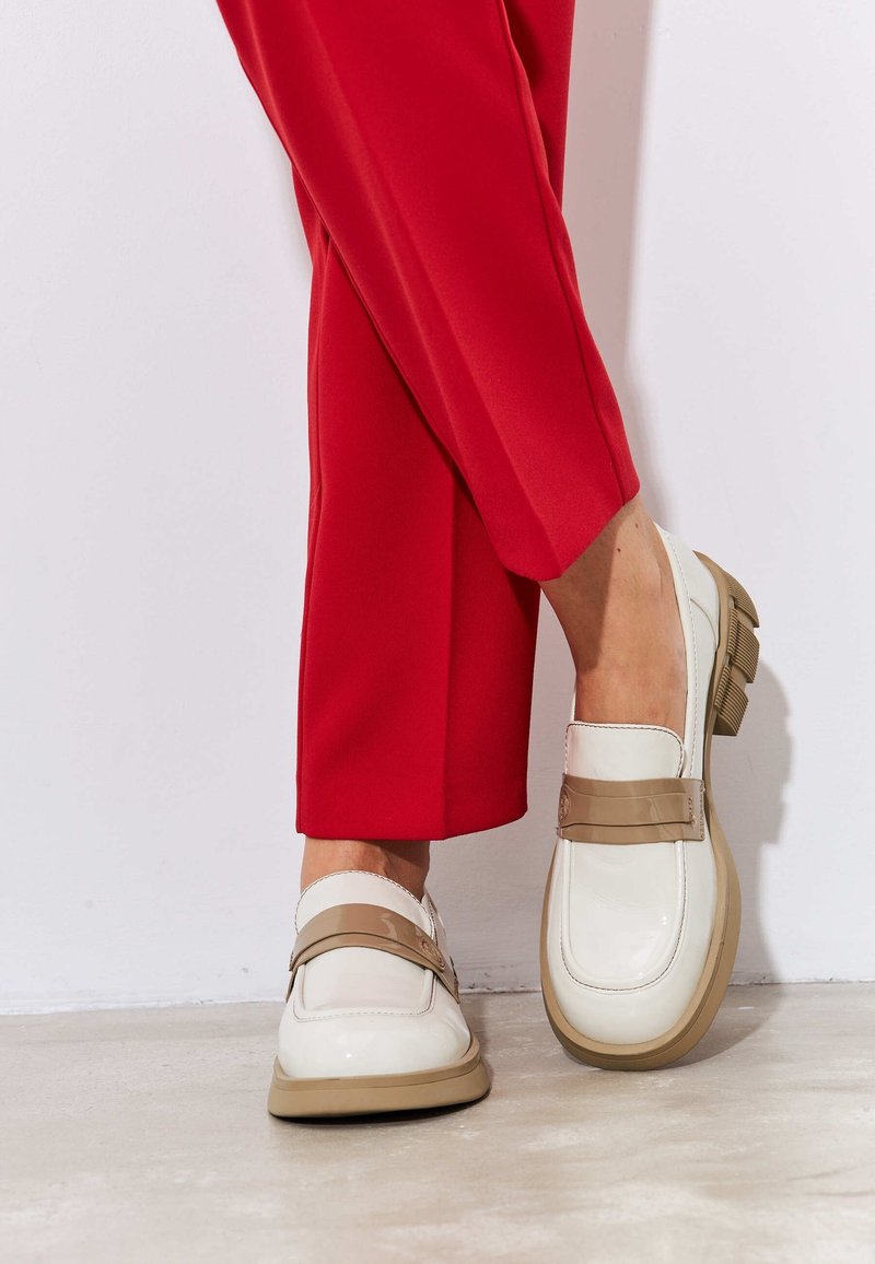 Personne portant des mocassins blancs brillants avec des brides et semelles beiges, assortis à un pantalon rouge vif, debout sur un sol clair.