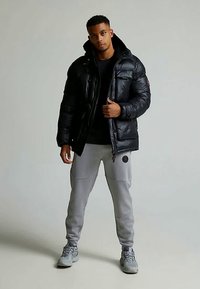 Svart vadderad jacka med huva, dragkedja framtill och två bröstfickor. Bärs med grå mjukisbyxor och grå sneakers. Slät textur.