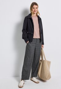 Giacca blu scuro con colletto, camicia rosa, pantaloni larghi grigi a quadri, e una borsa a tracolla beige texturizzata. Le sneakers sono color crema con dettagli marroni.