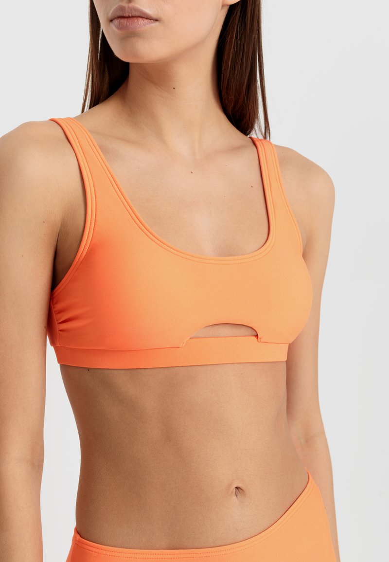 Oranžni športni modrček iz gladkega materiala, ki ima široke naramnice in osrednjo izrezano podrobnost, zasnovan za udobje in aktiven kroj.