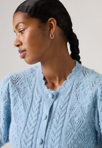 Mujer con cabello trenzado que lleva pequeños pendientes de aro y un cárdigan tejido azul claro con patrones texturizados y botones forrados de tela.