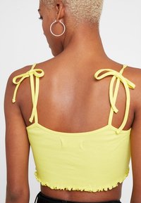 Top corto amarillo con textura acanalada, tirantes ajustables y dobladillo con volantes. La tela tiene una textura suave que resalta su diseño ajustado.