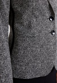 Veste ajustée en motif pied-de-poule noir et blanc, avec un col à revers cranté, trois boutons noirs et une poche latérale.