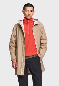 Homme portant un imperméable beige par-dessus une polaire orange zippée et un pantalon noir, debout avec une main dans la poche devant un fond uni.