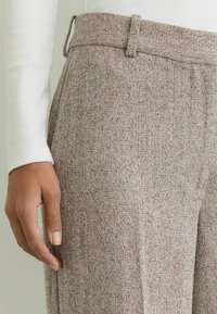 Püksid kalasaba mustriga, mis on valmistatud tekstuurse beeži kangaga, millel on peened pruunid täpid. Lai vöökoht ja küljetaskud on nähtavad.