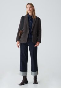 Blazer marrone con texture sopra una camicia blu con bottoni, abbinato a jeans a gamba larga in denim scuro con risvolti chiari e stivaletti marroni.