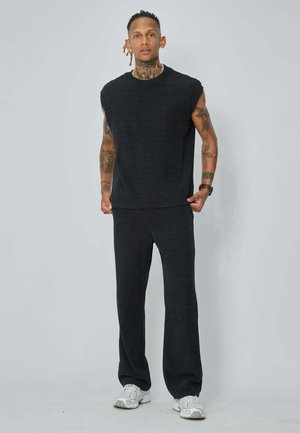 Homme avec des tatouages portant un haut noir sans manches et un pantalon avec des baskets blanches, debout devant un fond clair uni.