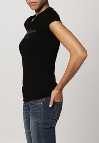 Henkilö, joka on pukeutunut tiukasti istuvaan mustaan lyhythihaiseen t-paitaan ja sinisiin farkkuihin, käsi takataskussa, kuvattu sivusta.