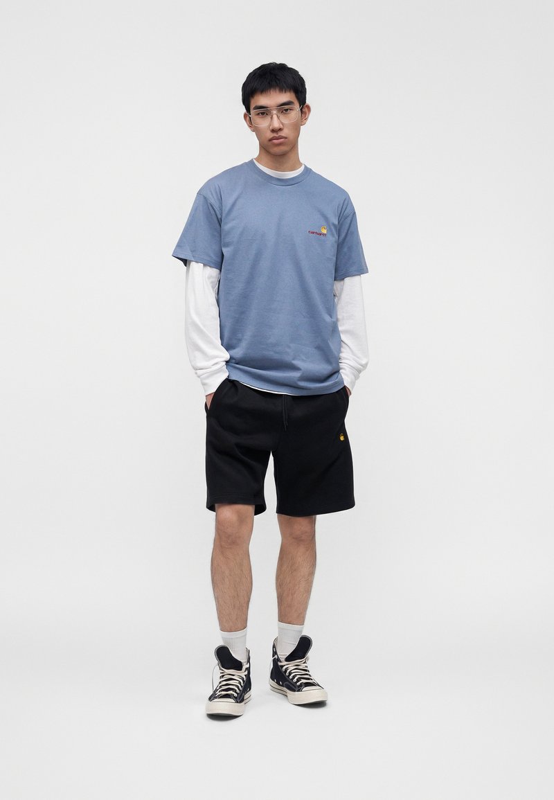 Young man standing with hands in pockets, wearing glasses, layered blue and white shirts, black shorts, white socks, and black high-top sneakers.
