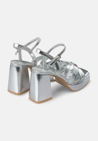 Sandalias de tacón alto de metal plateado con un grueso tacón cuadrado, diseño de tiras cruzadas y correa de tobillo ajustable. Textura suave y acabado brillante.