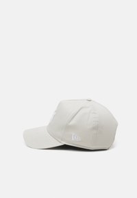 Beige Kappe mit einem gebogenen Schirm, mit einem weißen gestickten Logo auf der Vorderseite und einem kleinen weißen Akzent an der Seite.