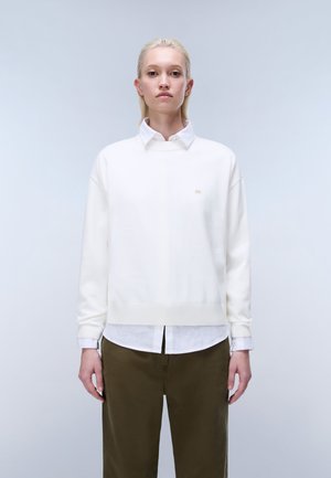 Jonge persoon met recht blond haar draagt een witte trui over een wit overhemd met kraag en olijfgroene broek, staand tegen een lichte achtergrond.