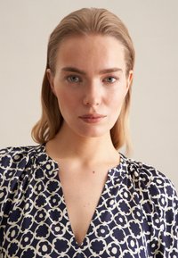 Granatowa bluzka w kwiatowy wzór z dekoltem w serek, krótkimi rękawami i plisowanymi detalami na ramionach, wykonana z lekkiego materiału.