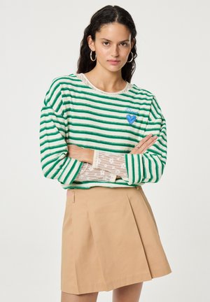 Vrouw met lang donker haar die een groen-wit gestreepte trui draagt met een blauwe hartpatch, kanten mouwen, een beige geplooide rok en oorringen.