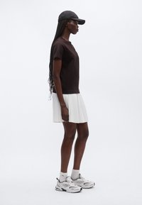 Brūns īsroku T-krekls, balta krokota svārki, balti sporta apavi ar acs detaļām un melna cepure, skatīts no sāniem.