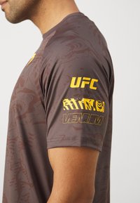 Kratke rokave, rjava športna majica z teksturiranim vzorcem, z rumenimi logotipi UFC in Venum na rokavih.