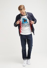 Marineblauer gesteppter Mantel mit weißem Besatz, rotem Innenfutter; weißes T-Shirt mit Cookie Monster Grafik; dunkle Jeans; weiße Sneakers.