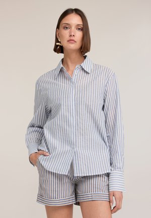 Frau trägt ein blau-braun vertikal gestreiftes Langarmshirt und passende gestreifte Shorts, steht mit einer Hand in der Tasche.