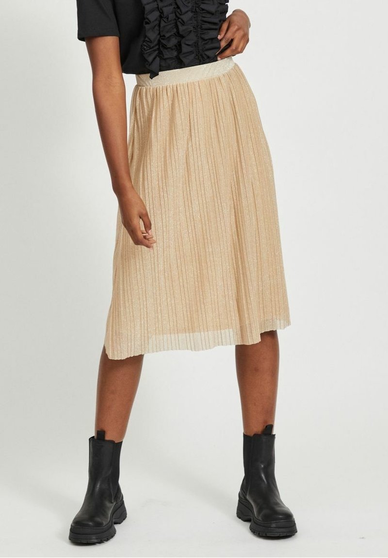 Femme portant une jupe midi plissée beige, un haut noir à volants et des bottines noires, debout devant un fond clair uni.