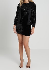 Černá flitrová bunda přes krátké černé šaty, které mají kombinaci texturovaných látek a hladký pásek. Jednoduché černé sandály na tenkém podpatku.