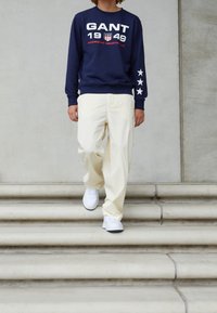 Marineblauwe sweatshirt met witte tekst en steraccenten, crèmekleurige broek en witte sneakers. Modell die stenen trap afdaalt.
